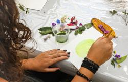 Com foco nas mãos de uma mulher a foto mostra uma mesa branca com flores e folhas em cima, um pote de tinta e uma pintura sendo feita pela mulher. 