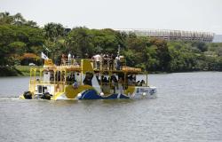 Foto de um barco com tonalidade amarela, azul e branca tansportando pessoas em navegação por espelho d'água, com o Mineirão ao fundo
