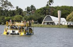 A imagem mostra o Capivarã, embarcação turística de cores branca e amarela, navegando pela Lagoa da Pampulha, em Belo Horizonte. Há passageiros a bordo, distribuídos nos dois pavimentos do barco. Ao fundo, aparece a Igreja São Francisco de Assis, um dos principais marcos arquitetônicos do Conjunto Moderno da Pampulha, cercada por árvores e palmeiras. O dia está claro, com céu parcialmente nublado, e a paisagem destaca a integração entre a lagoa, a vegetação e a arquitetura modernista.