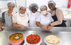 Cinco mulheres de avental e touca em uma cozinha com três tortas doces na mesa. 