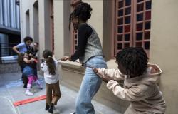 Crianças e adultos participam de uma brincadeira com uma corda vermelha em um corredor externo. Em primeiro plano, à direita, uma criança de cabelos crespos e conjunto bege segura a corda com as duas mãos e inclina o corpo para trás. Ao centro, uma adulta de tranças presas, colete xadrez e calça jeans também segura a corda. Outras participantes aparecem ao fundo. Há desenhos de giz no piso e janelas rosadas com vidros coloridos na parede.