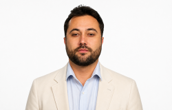 Homem de pele clara, com cabelo curto e escuro e barba cheia aparada, olhando diretamente para a câmera com expressão séria e neutra. Ele veste camisa social azul-clara e blazer bege claro. A foto tem fundo branco e iluminação suave, em estilo de retrato profissional.