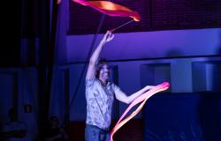  Em um palco sob iluminação azul, um artista circense sorri enquanto movimenta duas fitas coloridas que se desenrolam no ar. Ele segura cada fita por uma haste