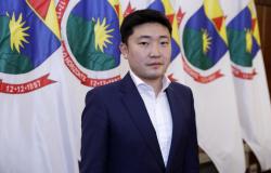 Cheng Qu, Secretário de Relações Institucionais, de terno, olhando em direção a câmera, e ao fundo, bandeiras de Belo Horizonte 