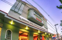 Fotografia em ângulo baixo da fachada do Cine Santa Tereza, um prédio histórico. No andar superior, há uma varanda curva sob pequenas janelas com o letreiro dourado "CINE SANTA TEREZA" iluminado. Luzes quentes embutidas na marquise iluminam a calçada, enquanto cabos elétricos cruzam o céu azul ao fundo.