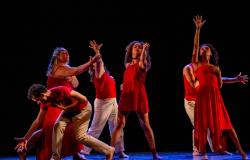 Cena do espetáculo de dança "Sangue Latino" em um palco escuro. Seis dançarinos descalços vestem figurinos em tom vermelho e branco. Com expressões dramáticas e mãos abertas, eles realizam movimentos expansivos: alguns em pé com os braços erguidos para o alto, e um homem agachado à esquerda inclinado sobre o chão.