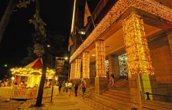 Fachada da Prefeitura Municipal de Belo Horizonte, com luzes natalinas e carrossel à frente, também iluminado, no início da noite. 