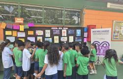 Grupo de crianças de uniforme escolar reunidas de costas, a observar uma exposição de desenhos e cartazes na parede sob o título "Galeria das Vozes".