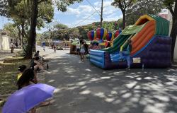  Em uma rua arborizada e fechada para o trânsito, acontece uma edição do programa "A Rua é Nossa". À direita, há grandes brinquedos infláveis coloridos, entre eles um escorregador nas cores verde, laranja, azul e roxo e estruturas em formato de balões. Crianças e adultos circulam e brincam pelo espaço. Algumas pessoas acompanham as atividades em pé, enquanto outras estão sentadas à sombra das árvores, na calçada à esquerda. Em primeiro plano, uma pessoa se protege do sol com uma sombrinha roxa. Ao fundo, ap