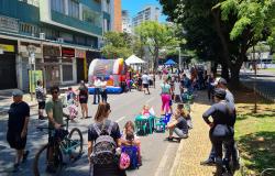 Em uma rua fechada ao trânsito, durante um dia ensolarado, crianças e adultos participam de atividades de lazer. Ao centro, há brinquedos infláveis coloridos, tendas, pequenas mesas e cadeiras infantis. Algumas crianças brincam, enquanto outras pessoas caminham, conversam ou acompanham as atividades. Em primeiro plano, há pessoas com bicicletas e uma mulher com mochila próxima a um grupo de crianças. À direita, árvores grandes fazem sombra sobre a calçada, onde outros participantes estão reunidos. Ao fundo,