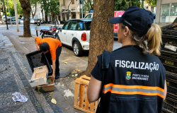 Fiscal da PBH, de costas, usando colete da Fiscalização e boné, aparece em primeiro plano, com homem recolhendo resíduos ao fundo.
