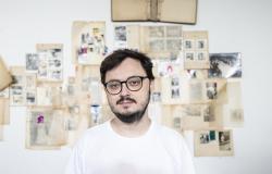 Imagem do artista Benedito Ferreira, um homem de óculos de armação preta e camiseta branca, de barba e bigode, posando diante de uma parede coberta por fotografias, documentos e imagens antigas organizadas em painéis.