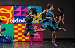 Fotografia horizontal de uma apresentação de dança em um palco com piso preto e brilhante. Três bailarinas executam movimentos coreografados. Em primeiro plano, à direita, uma bailarina de cabelos curtos, blusa e saia-calça azul-petróleo, com tatuagens nos braços, realiza um salto com uma das pernas estendida para trás. À esquerda, outra bailarina, de cabelos presos, veste blusa escura e calça larga, com um dos braços flexionado à frente do corpo. Ao fundo, à direita, a terceira bailarina, de blusa preta e 
