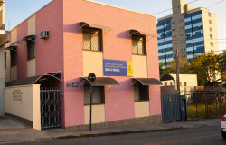 A imagem mostra a sede do Centro Especializado de Atendimento à Mulher. Trata-se de uma casa de dois andares com fachada predominantemente rosa, de tijolos aparentes. A parte inferior das paredes é revestida com pedras em tons de cinza. Na fachada há várias janelas protegidas por grades metálicas e cobertas por pequenas marquises curvas de metal. No centro, há uma placa azul e amarela com a identificação “Centro Especializado de Atendimento à Mulher Benvinda”, acompanhada da marca da Prefeitura de Belo Hor