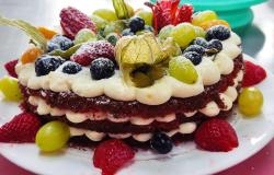 Imagem mostra um bolo Red Velvet inteiro, coberto por creme e frutas vermelhas e amarelas. O bolo está disposto em um prato branco.