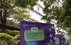 Placa roxa em primeiro plano exibe o texto "Educação EJA Escola Municipal Caio Líbano Soares" junto ao brasão da Prefeitura de Belo Horizonte. Ao lado, há outra placa similar. Ao fundo, vê-se a fachada de um prédio claro de vários andares com janelas e detalhes em azul, cercado pelas folhagens e galhos verdes de árvores frondosas que emolduram a parte superior da imagem.