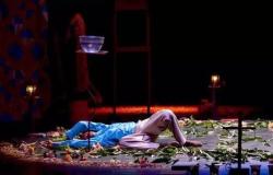 A imagem mostra um palco com fundo escuro e iluminação suave destacando a cena dramática, ao centro. Um homem negro está deitado de costas sobre o chão escuro, cercado por flores e folhas verdes espalhadas. Ele veste uma camisa azul de mangas compridas, calças claras e está descalço