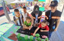 Foto de um evento comunitário ao ar livre, com quatro crianças participando de uma atividade em uma mesa, acompanhadas por duas servidoras uniformizadas. Sobre a mesa, há uma maquete com árvores, casas e veículos de brinquedo. Ao fundo, aparecem tendas, pessoas e materiais do evento.