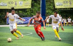 Em um campo de futebol society com gramado sintético, três jogadores disputam a bola. À esquerda, um atleta de uniforme branco, com detalhes dourados e meias amarelas, corre em direção à bola, que está à sua frente. No centro, um jogador com camisa cinza e laranja, calção vermelho e meias vermelhas acompanha a jogada. À direita, outro atleta de branco e amarelo também corre para alcançar a bola. Ao fundo, há jogadores reservas sentados no banco, alambrados, árvores e faixas de publicidade.