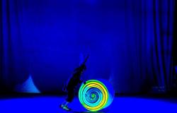 Em um palco iluminado por uma forte luz azul monocromática, um artista caracterizado empurra um grande cilindro decorado com uma espiral luminosa em tons de amarelo e verde. O performer veste casaco escuro, calças médias com meias listradas e um chapéu adornado com grandes penas. O fundo é composto por cortinas escuras sob a iluminação azul.
