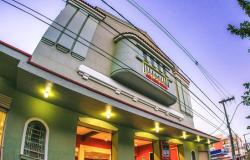 Fotografia em ângulo baixo da fachada do Cine Santa Tereza ao entardecer, prédio histórico em estilo Art Déco, em tom verde-claro, com frisos geométricos e um balcão curvado no centro, onde se destaca o letreiro vermelho e dourado com o nome do cinema.