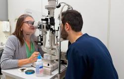 Médica oftalmologista vestida com blusa verde e com casaco cinza atende um paciente de barba vestido com uma blusa azul de mangas compridas.