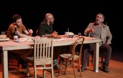 Três atores em cena de teatro conversam ao redor de uma mesa de madeira com livros, garrafa de vinho e copos; à direita, um homem mais velho aponta o dedo enquanto fala, sob fundo completamente escuro.