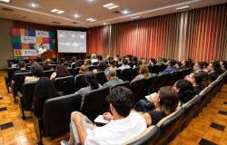 A imagem mostra, do fundo de um auditório iluminado, uma plateia de costas atenta a uma palestra. No palco, à esquerda, um palestrante fala num púlpito junto a um painel colorido com a inscrição "Rio de Histórias - Encontro de Legislação". Ao centro, uma tela exibe slides com textos e imagens, e a lateral do recinto está coberta por uma cortina terracota.
