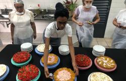 Imagem mostra quatro mulheres vestidas com dolmãs em frente ao mesa com várias tortas. Uma delas está manipulando uma das tortas enquanto as outras observam.