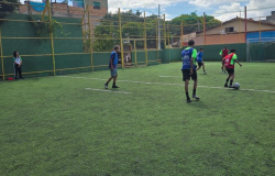 Jovens usando coletes azuis e vermelhos jogando futebol em um campo de gramado, enquanto professora observa a partida