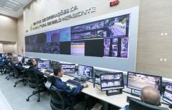 Cinco agentes da Guarda Municipal de BH sentados em frente a monitores de computador na mesa e um painel maior na parede que mostram imagens de diferentes locais de Belo Horizonte 