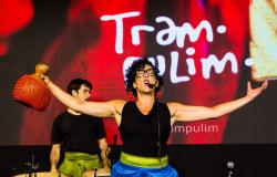 Uma artista no centro do palco expressa grande emoção ao cantar com a boca aberta e braços bem abertos. Ela tem cabelos cacheados pretos, usa óculos de grau, brincos grandes e blusa preta sem mangas. Em uma das mãos, segura um instrumento percussivo (xequerê) para o alto. Ao fundo, à esquerda, um homem de camiseta preta toca tambores. Atrás deles, um telão iluminado em tom vermelho exibe a palavra "Trampolim" escrita em letras brancas estilizadas.