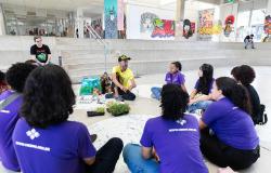 Nove jovens e um educador posam agachados em círculo, segurando pequenas mudas de plantas. No centro, há bandejas com mudas, cartelas de ovos e materiais utilizados na atividade. Ao fundo, o espaço do CRJ exibe murais coloridos e um auditório.