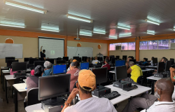 Sala de aula equipada com computadores. Professor em frente ao quadro e alunos sentados de frente para computadores, assistindo a aula.