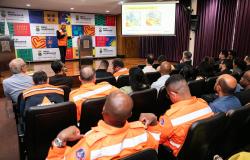 Imagem mostra várias pessoas uniformizadas com coletes laranja assistindo a uma apresentação de um homem tambémn de colete laranja. No fundo há um banner com logomarcas da PBH.