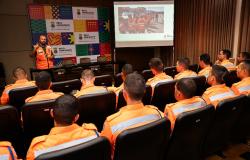 Imagem mostra uma pessoa da Defesa Civil, vestoda com uma colete laranja em uma cenário com uma apresentação de power point atrás dele. Há também uma banner com identidade visual da Prefeitura. Na platéia há vários oficiais dos Bombeiros assistindo à palestra. Todos estão com uniformes laranja do coorporação.