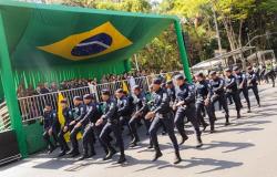 Guardas Municipais marchando em formação em frente a um palco coberto com uma grande bandeira brasileira pendurada atrás deles. O palco estão autoridades e oficiais.