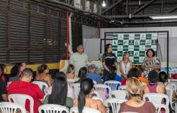 Imagem mostra uma sala com várias pessoas de costas assistindo a uma aula. Há três pessoas de pé e uma assentada. Todas estão em frente a um banner.