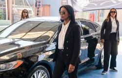 Uma mulher caminha usando óculos escuros, camisa branca e terno preto. Ao lado dela, há um carro estacionado em uma área coberta. Ao fundo e à direita, outras duas mulheres vestidas com trajes semelhantes e óculos escuros mantêm uma postura atenta.