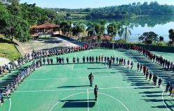 Grupo de jovens e adultos do projeto de escotismo vestindo uniformes são organizados em formato de "U" sobre uma quadra poliesportiva ao ar livre. Ao fundo, destaca-se uma paisagem natural, com um lago sob um céu azul sem nuvens.