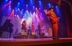Em um palco iluminado por luzes azuis, roxas, vermelhas e amarelas, um artista negro ocupa o primeiro plano, à direita, usando terno xadrez vermelho, chapéu de aba larga e sapatos escuros. Ao fundo, três vocalistas cantam com microfones, acompanhados por músicos e instrumentos, entre eles bateria, baixo e um piano de cauda à esquerda.
