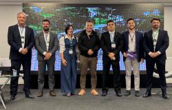 Grupo de sete pessoas, sendo seis homens e uma mulher, alinhadas lado a lado num palco, durante o evento. Ao fundo, encontra-se painel de LED que exibe uma imagem de uma paisagem natural com água e vegetação.