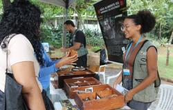 Mulher usando colete da Prefeitura de Belo Horizonte interagindo com visitantes do parque, onde caixas com sementes e materiais naturais estão expostos, ao lado de banner educativo sobre não alimentar animais silvestres