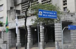 A imagem mostra a fachada do prédio sede da Prefeitura de Belo Horizonte, com duas placas onde está escrito Prefeitura de Belo Horizonte, mais uma bandeira do Brasil e uma bandeira de Belo Horizonte hasteadas; na calçada uma pedestre caminha em frente ao prédio.