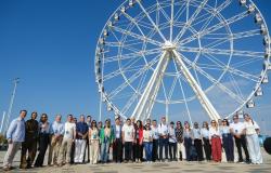 Representantes de diferentes cidades posam para foto coletiva em frente a uma grande roda-gigante. O grupo está reunido em uma área aberta, sob céu azul e tempo ensolarado. 