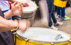 Uma imagem em plano médio e close-up foca em um percussionista tocando tambores amarelos. A ação é dinâmica, e suas mãos e as baquetas estão desfocadas para transmitir velocidade e movimento. Ele está segurando uma baqueta no tambor principal em primeiro plano, com o outro tambor e outras pessoas ao fundo.