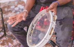 Imagem das mãos de um homem negro tocando um pandeiro transparente. A mão direita do homem, que usa uma pulseira branca, está em movimento, posicionada acima do instrumento. Dá para ver que o homem usa uma calça cinza escura.