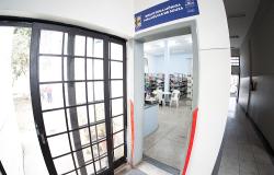 Fotografia em ângulo aberto da entrada da biblioteca. Uma porta cinza está aberta. Acima dela, há uma placa azul onde se lê "Biblioteca Mônica Malaquias de Souza". As fitas vermelhas de inauguração estão cortadas e penduradas nas laterais do batente. Pela abertura da porta, vê-se o interior com estantes de livros, mesa e cadeiras. À esquerda, há uma grande janela quadriculada, e à direita, um corredor longo.