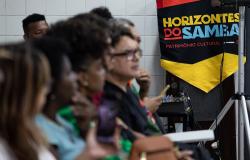  Fotografia colorida de um grupo de pessoas sentadas em fileiras, no canto esquerdo da imagem. Elas estão de perfil, olhando para a direita. As pessoas do primeiro plano estão desfocadas, enquanto o foco principal está em um homem de óculos no centro. No canto superior direito, há um banner vertical pendurado em um tripé de metal, com o design nas cores vermelho, preto e amarelo. No banner, lê-se o título "Horizontes do Samba". A imagem foi registrada durante a 2ª Reunião Pública, em 2025.