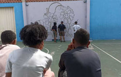 Três jovens sentados e observando outros jovens fazendo grafitti em um muro branco, a ilustração na parede é um conjunto de flores